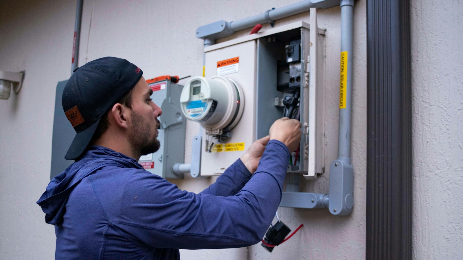 Monteur in blauwe hoodie werkt aan een elektriciteitsmeter, het soort werkdag dat je in een vacaturetekst beschrijft